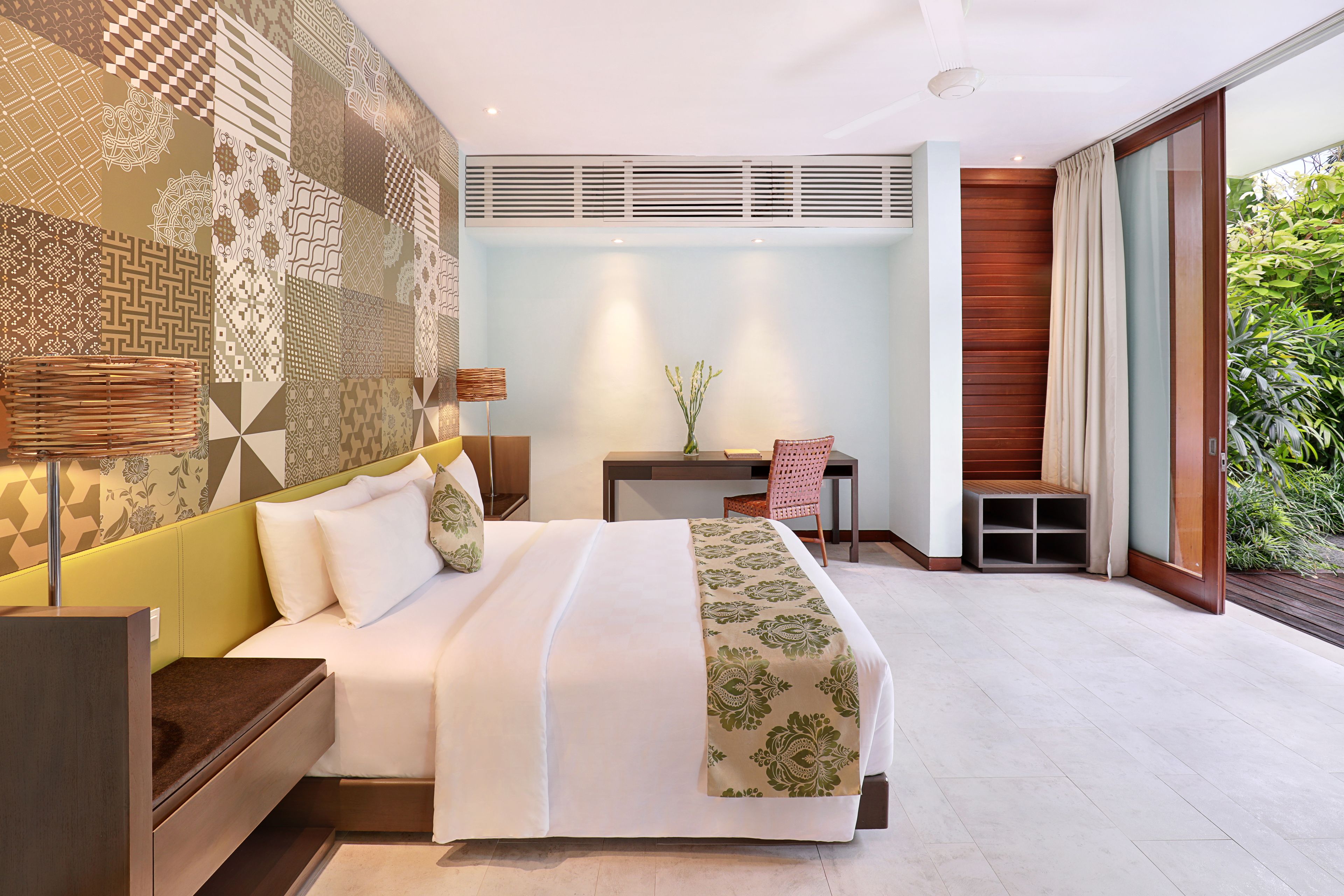 A modern hotel room with a large bed, a desk, and a chair. There is a small table with a vase of flowers and a window with curtains.