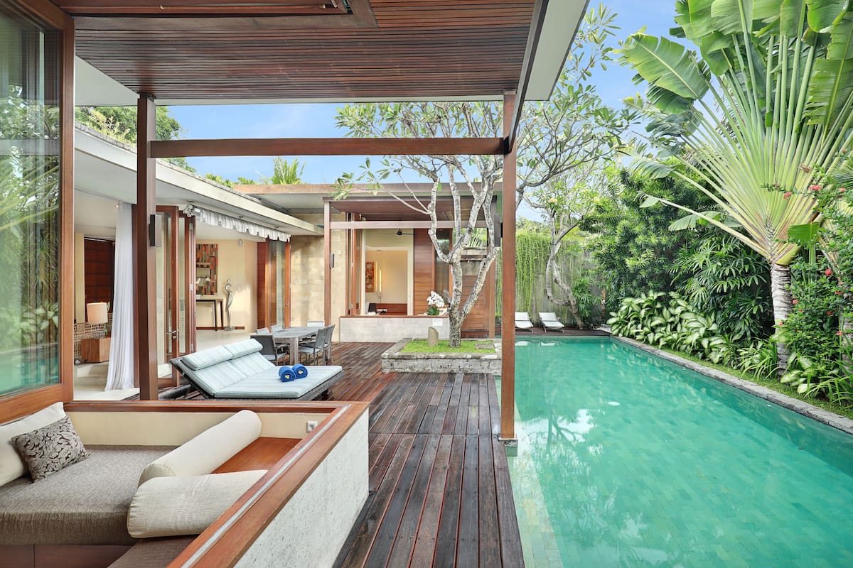 Uma área de piscina moderna ao ar livre, com deck de madeira, espaço de estar com almofadas e vegetação exuberante.