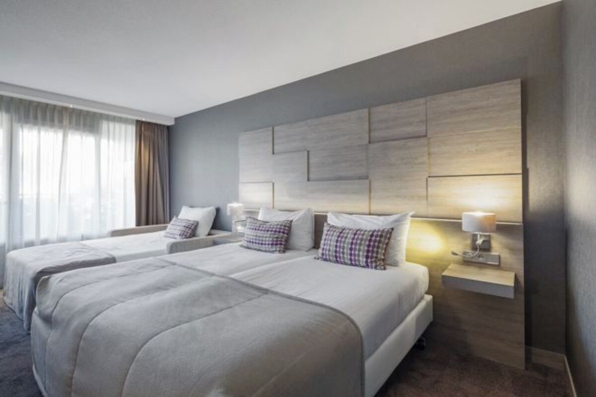 A modern hotel room with two beds, a large wall-mounted headboard, bedside lamps, and a view of the outdoors through a window with curtains.