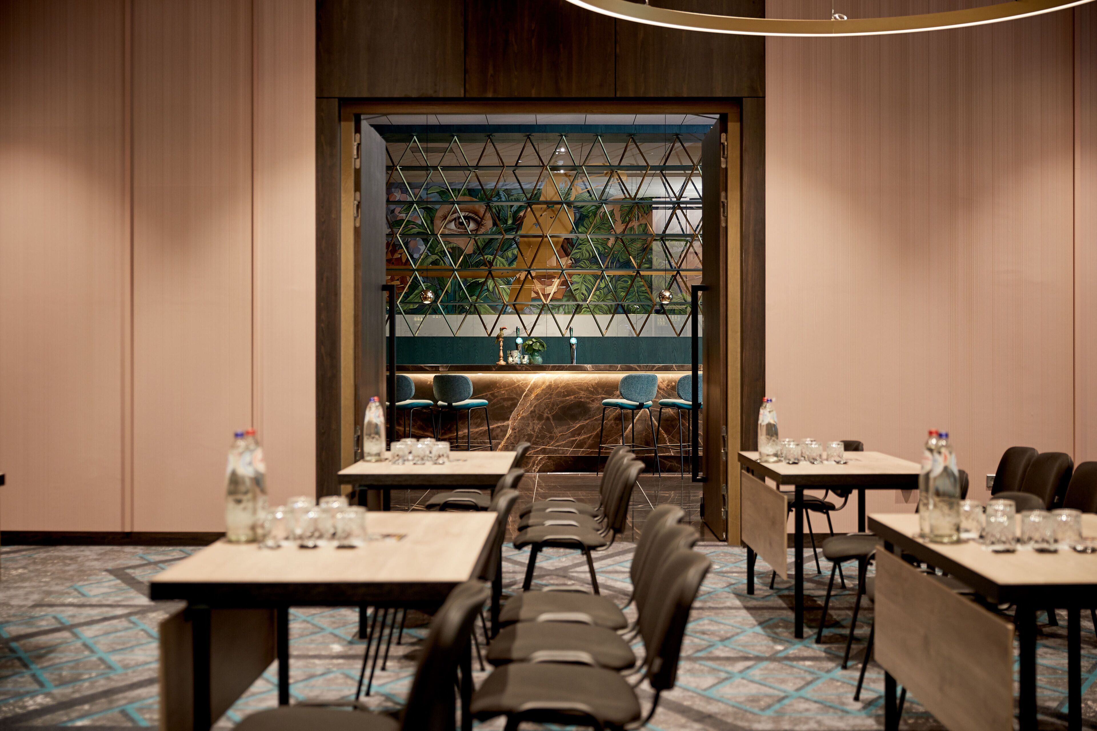 Une salle à manger d’hôtel avec des tables dressées pour les clients, un mur décoratif orné d’un motif géométrique et une vue sur l’extérieur à travers une fenêtre où l’on aperçoit une plante.