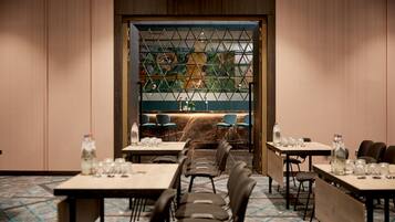 Une salle à manger d’hôtel avec des tables dressées pour les clients, un mur décoratif orné d’un motif géométrique et une vue sur l’extérieur à travers une fenêtre où l’on aperçoit une plante.