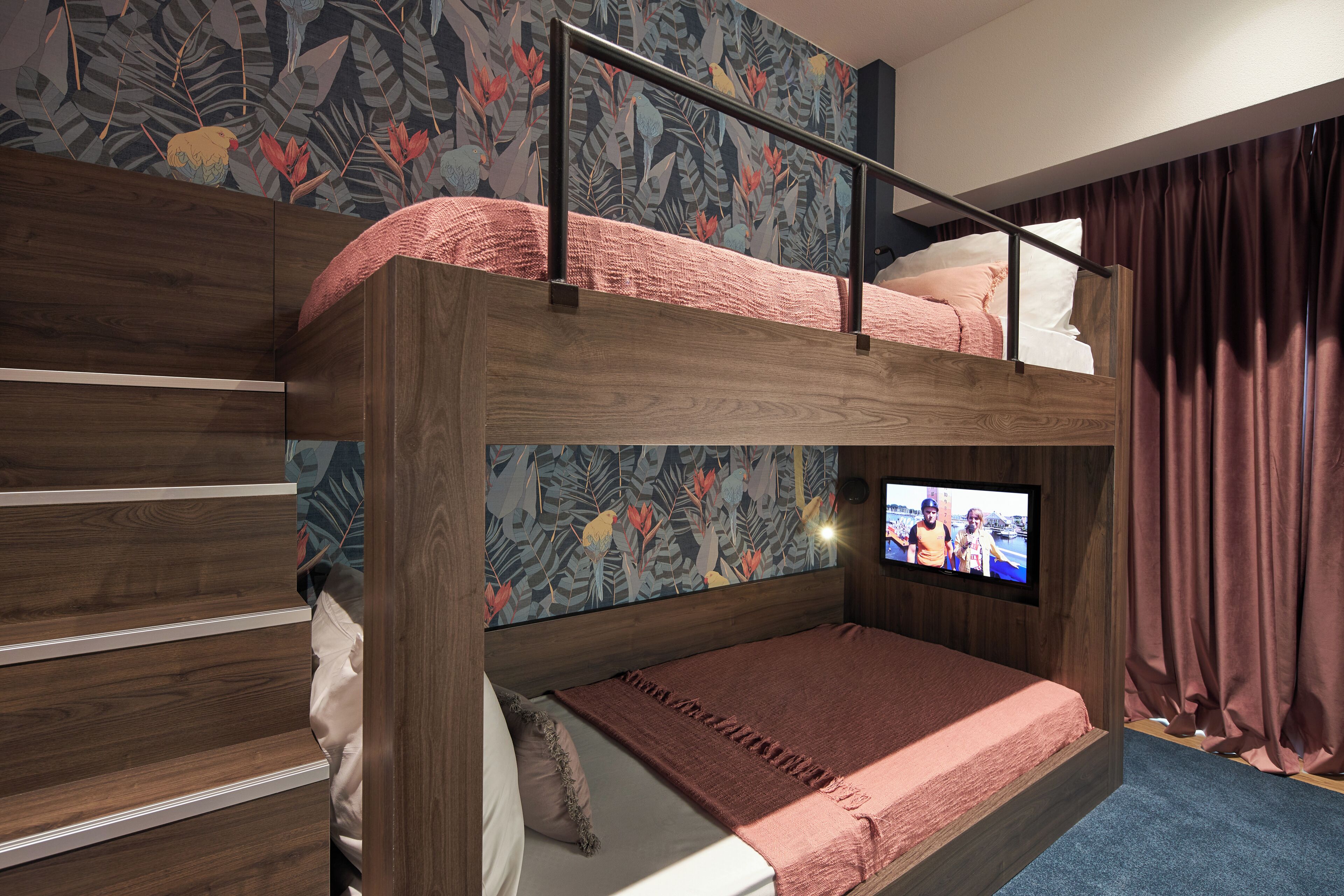 A bunk bed with a television on the lower bunk, a wooden headboard, and a floral wallpaper.