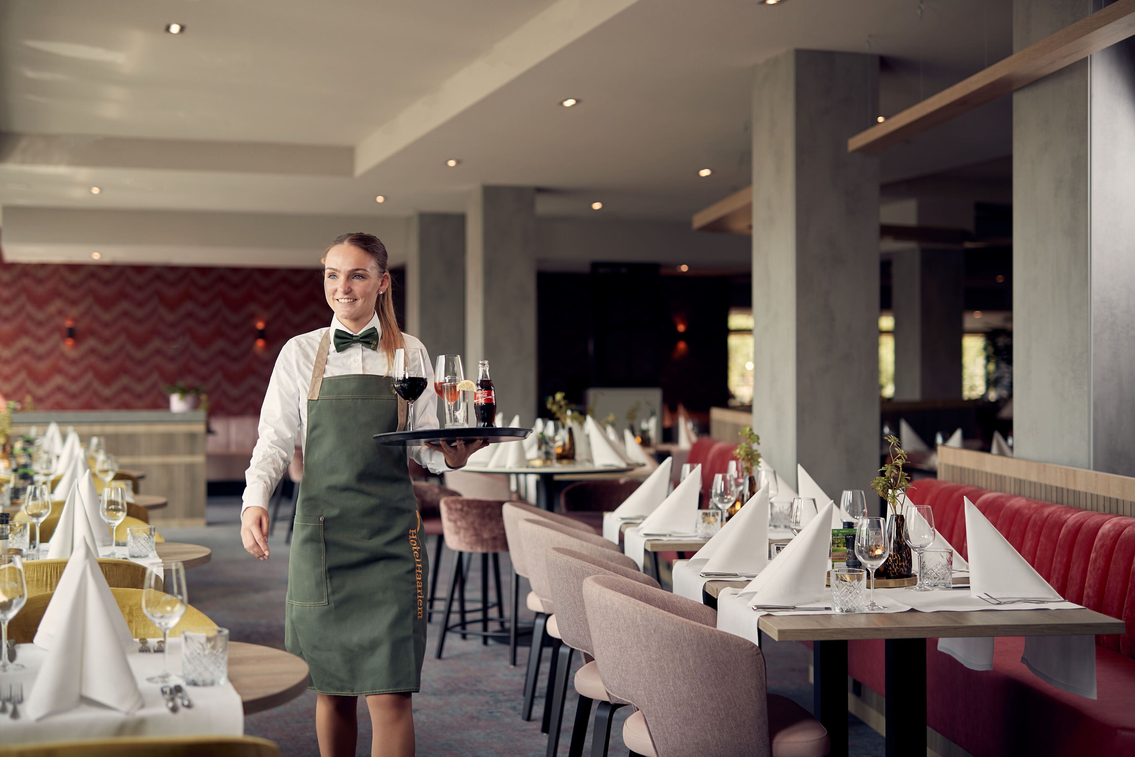 A restaurant with tables set for dining, a waiter carrying a tray, and a red upholstered booth in the background.