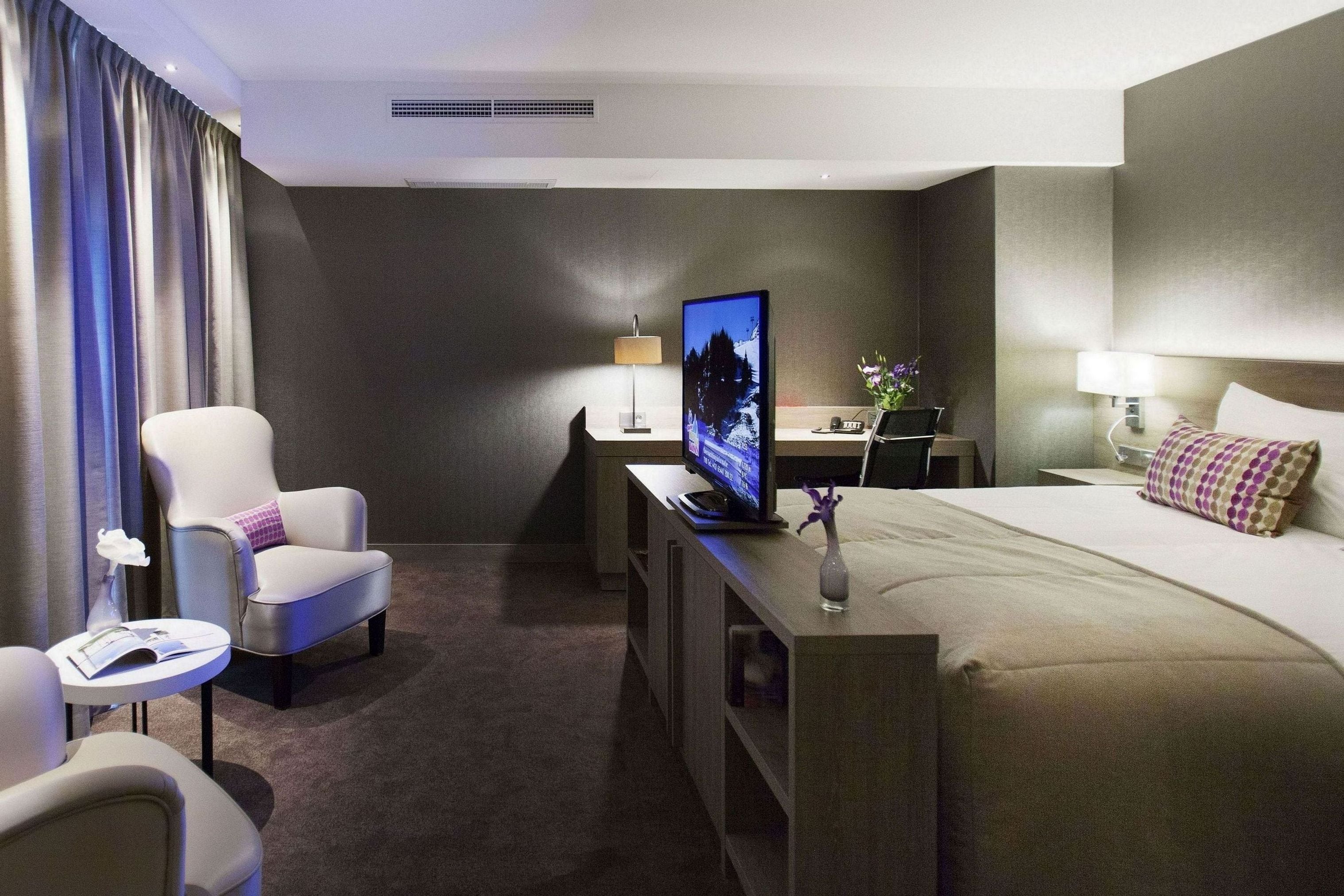 Una habitación de hotel con cama, escritorio con computadora, televisión, silla y mesita de noche con lámpara.