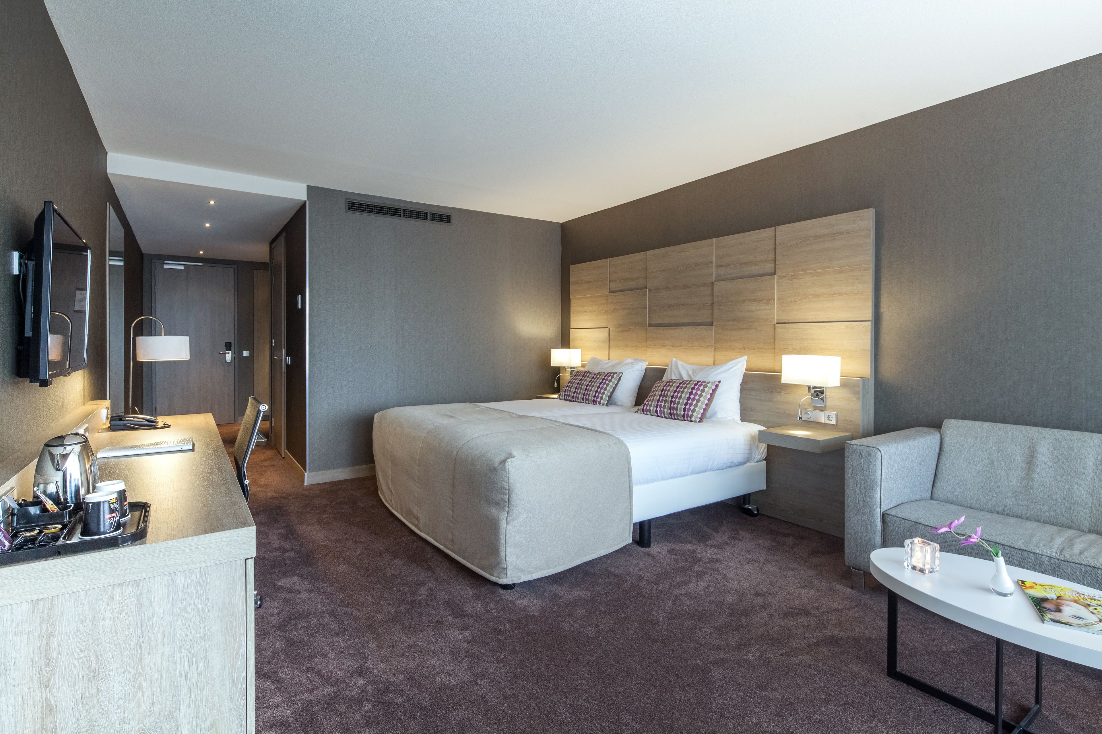A modern hotel room with a large bed, a sofa, a desk with a lamp, and a TV.