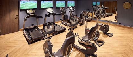 A gym with multiple cardio machines, a wooden floor, and large screens displaying sports events.