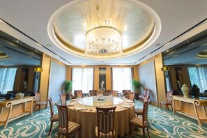 Un salón de conferencias de hotel con una mesa redonda, sillas, un candelabro y ventanas amplias.