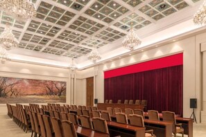 Un salón de conferencias con una larga mesa y sillas, un telón rojo de fondo y un techo decorado.