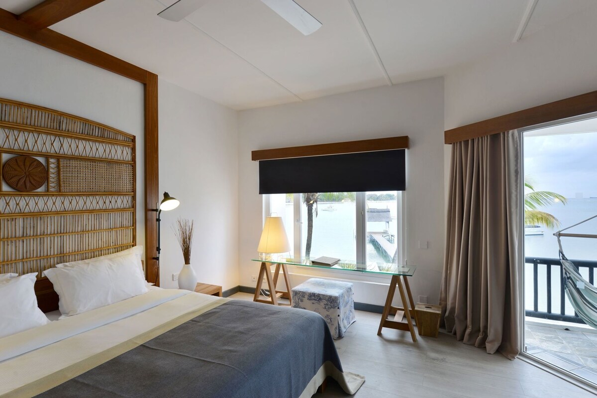 Une chambre d’hôtel avec un lit, une table de chevet, une lampe, une fenêtre avec des rideaux et une vue sur la mer.