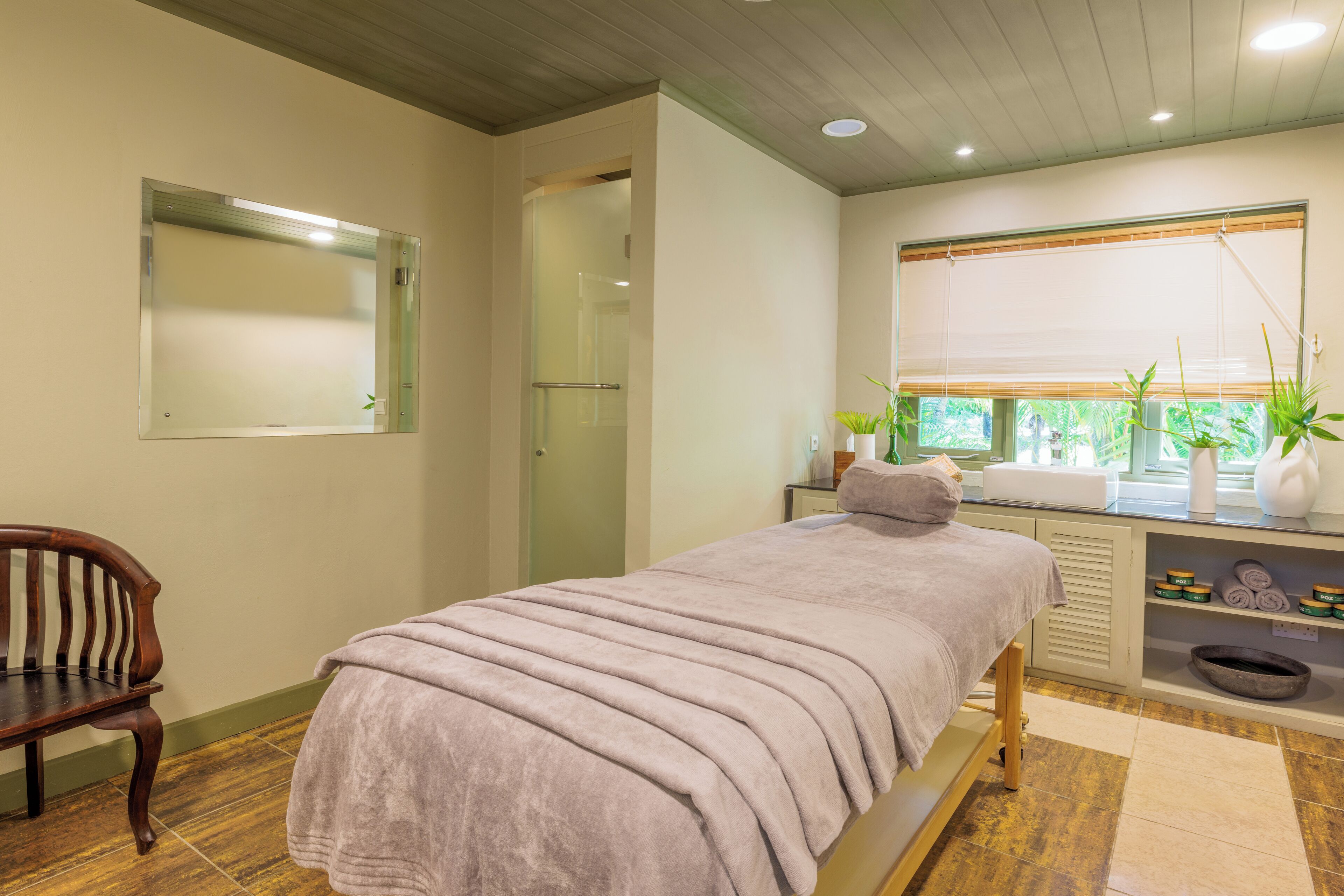 A room with a bed, a chair, a window with plants, and a shelf with towels and toiletries.