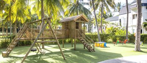 A playground with a wooden swing set and slide, surrounded by palm trees and a building in the background.