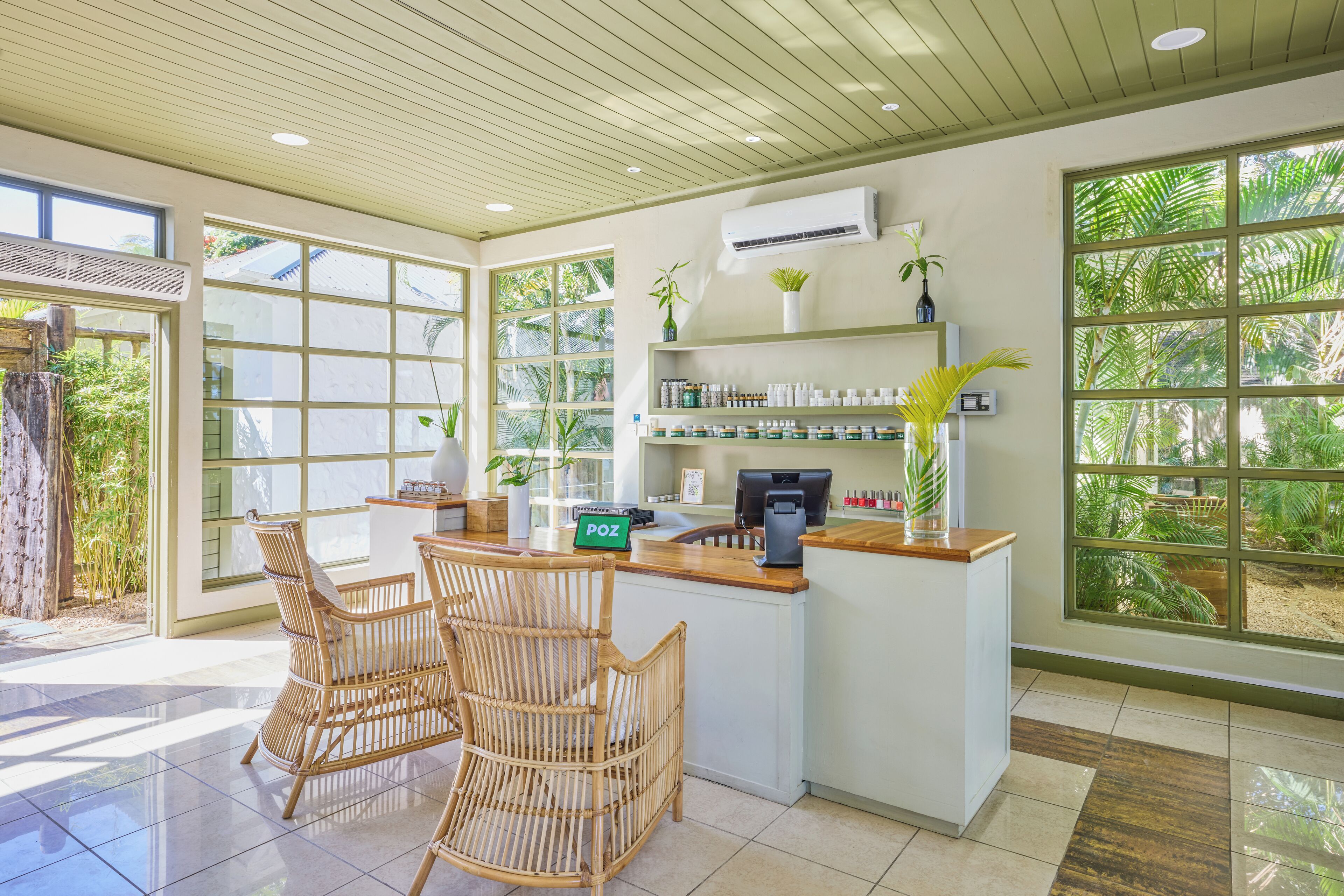 A reception area with wicker chairs, a counter with a computer, shelves with products, and large windows with a view of greenery.