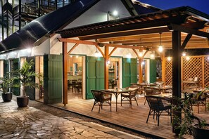 An outdoor patio with wooden tables and chairs, green shutters, and potted plants. - The Ravenala Attitude (Balaclava)