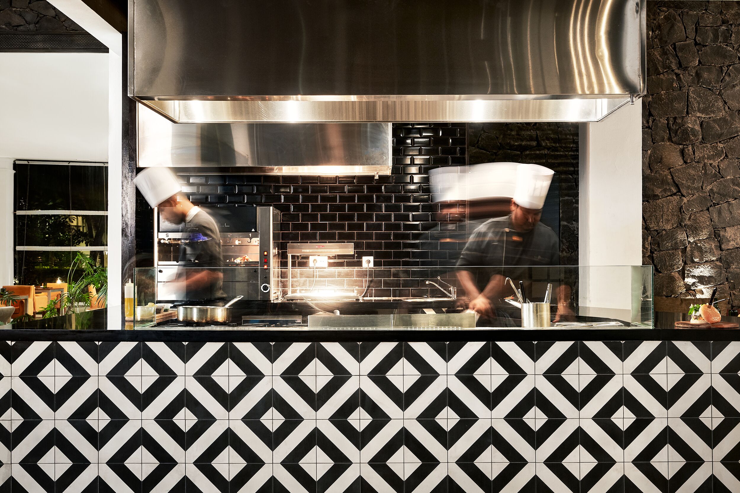 A modern kitchen with a stainless steel hood, black tiled backsplash, and a patterned tile floor.