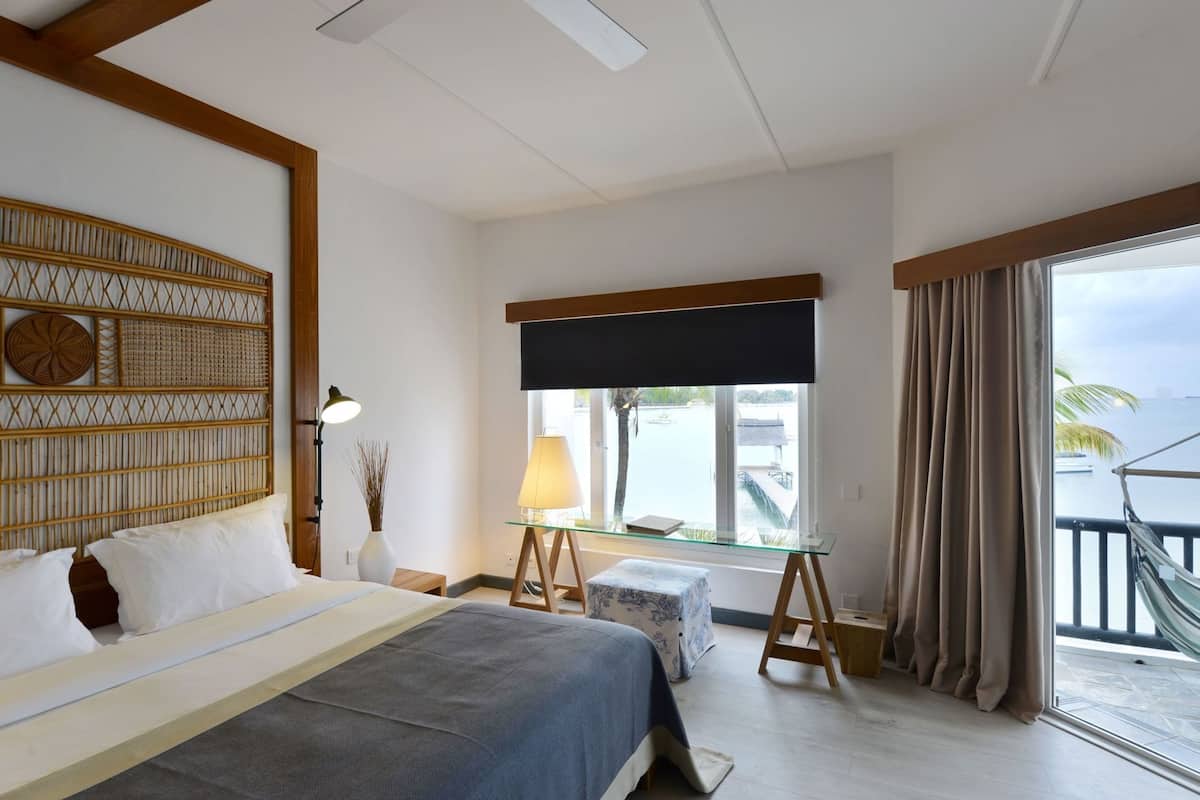 A hotel room with a bed, bedside table, lamp, window with curtains, and a view of the sea.
