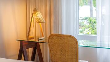A room with a bed, a wicker chair, a glass table with a lamp, and a window with curtains.