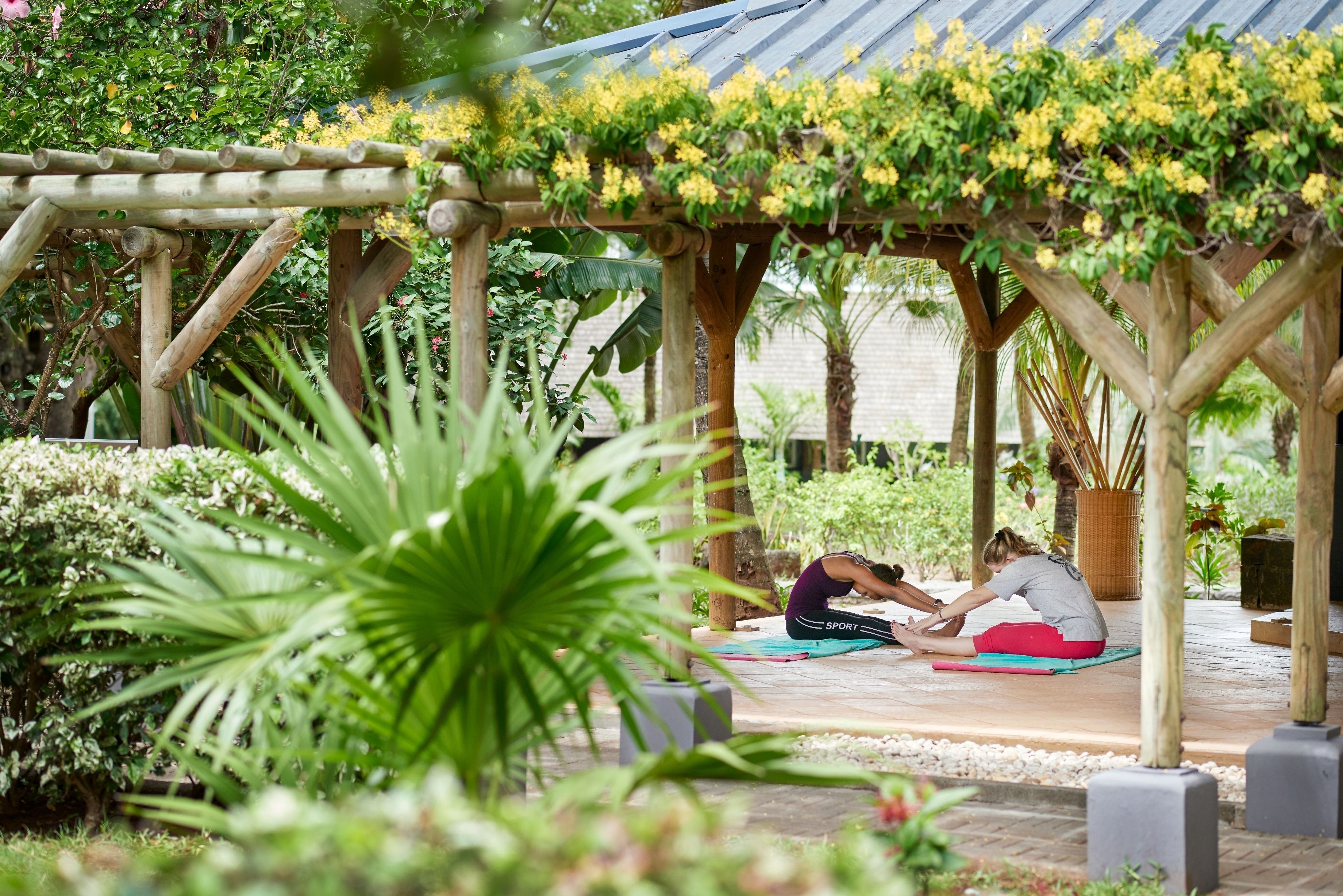 A wooden pergola with yellow flowering vines, two people exercising on mats, and lush greenery in a garden setting.