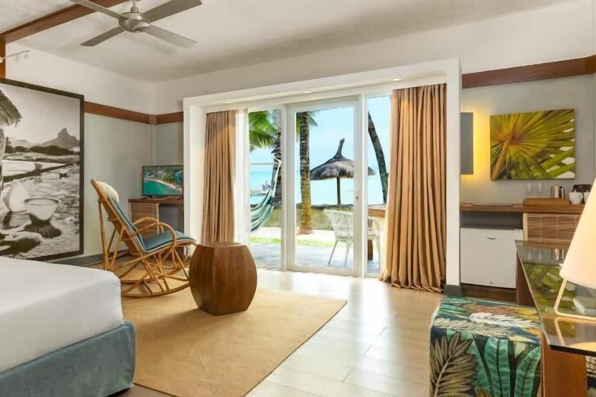 A hotel room with a bed, a chair, a table, a TV, and a view of the beach through the sliding doors.