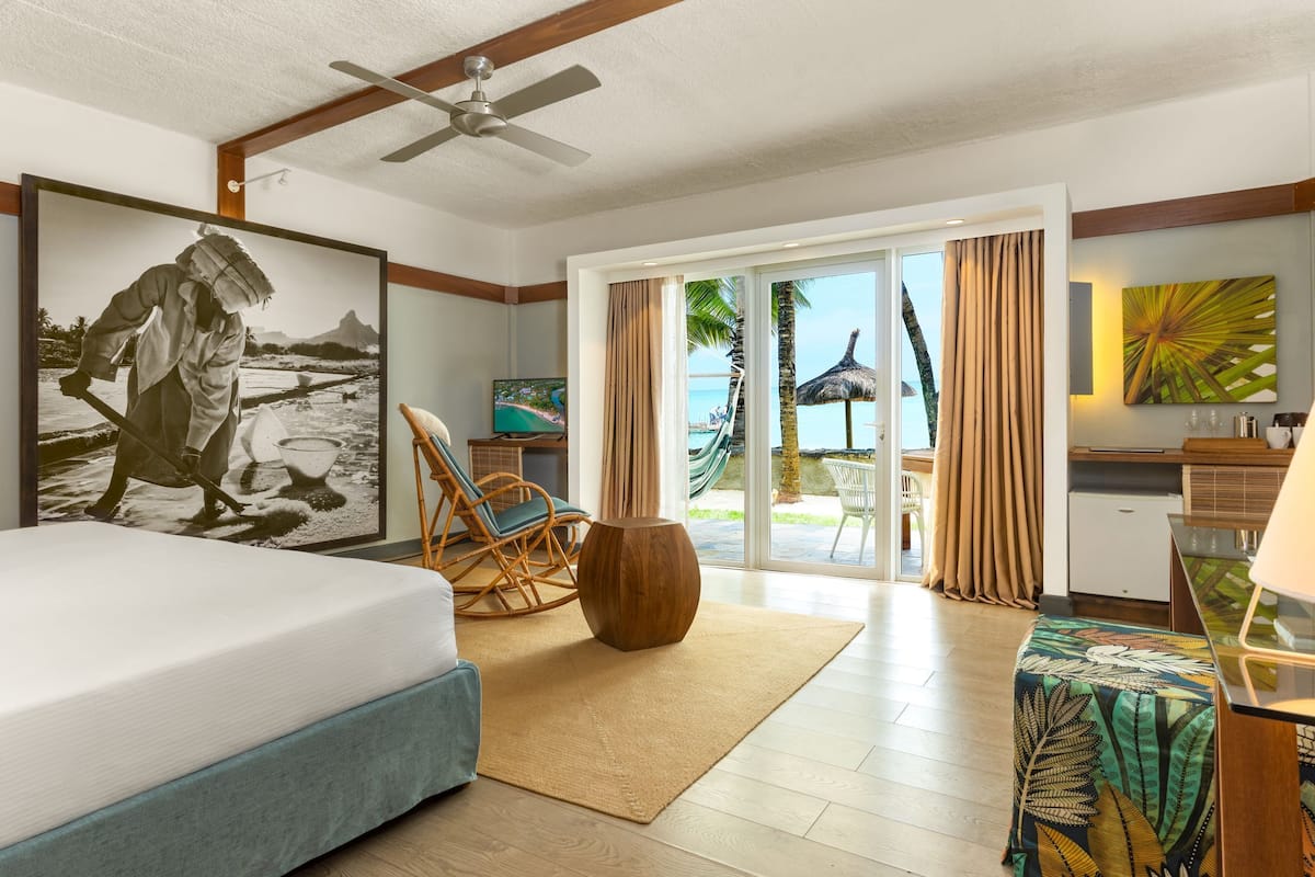 A hotel room with a bed, a wooden floor, a ceiling fan, a large window with curtains, a view of the beach, a dining area, and a painting on the wall.