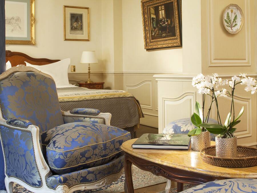 A room with a bed, a blue upholstered armchair, a small table with a book and orchid plants, and a nightstand with a lamp.