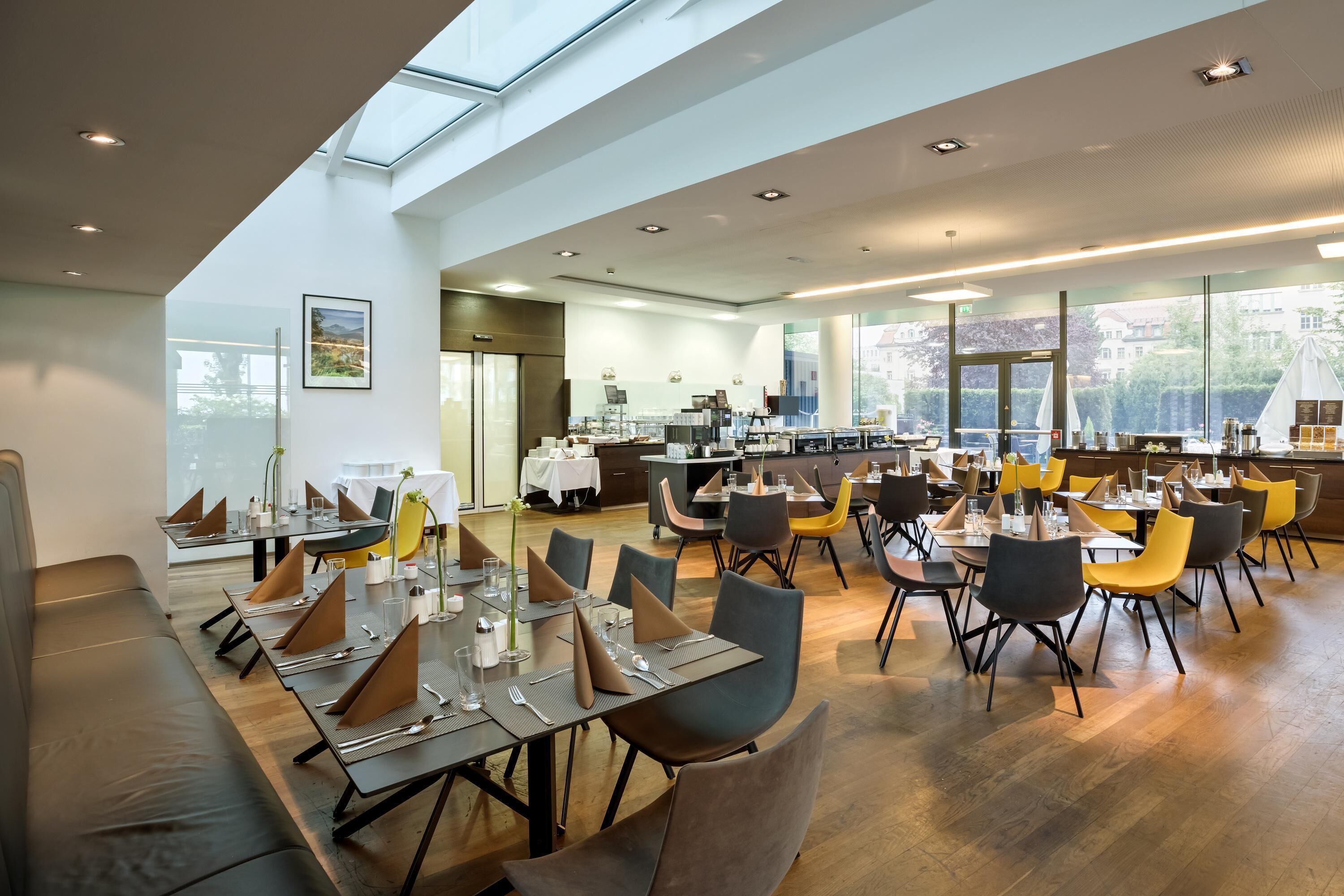 A modern dining area with long tables, chairs, and a kitchen in the background.