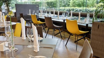 A restaurant with tables set for dining, featuring a mix of yellow and brown chairs, and a view of greenery outside.
