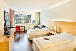 A hotel room with two beds, a desk, a TV, and a window with a view of buildings. - Austria Trend Hotel Congress Innsbruck (Innsbruck)