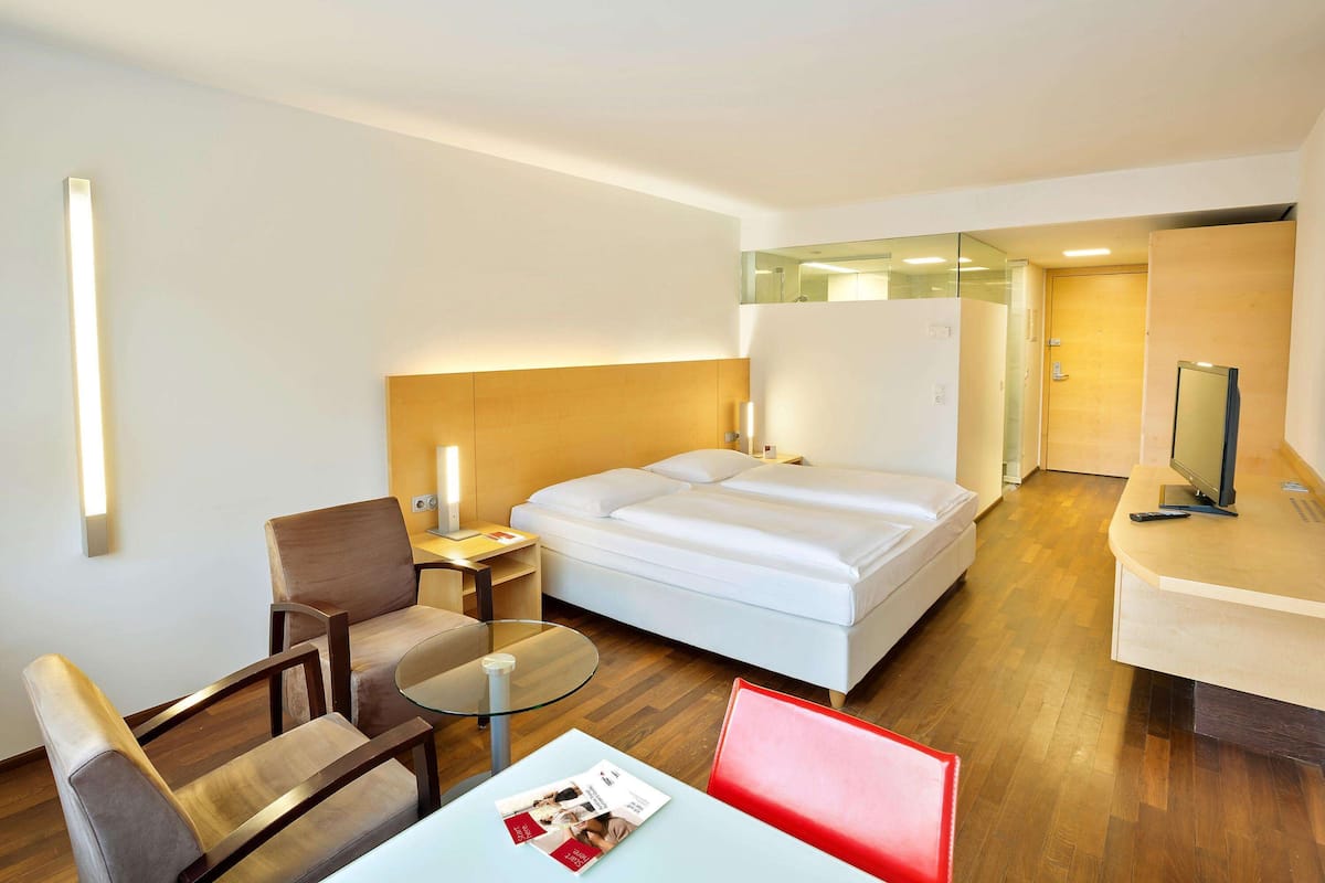 Una camera d'albergo con un letto, una scrivania, una sedia, un tavolino e un divisorio in vetro.