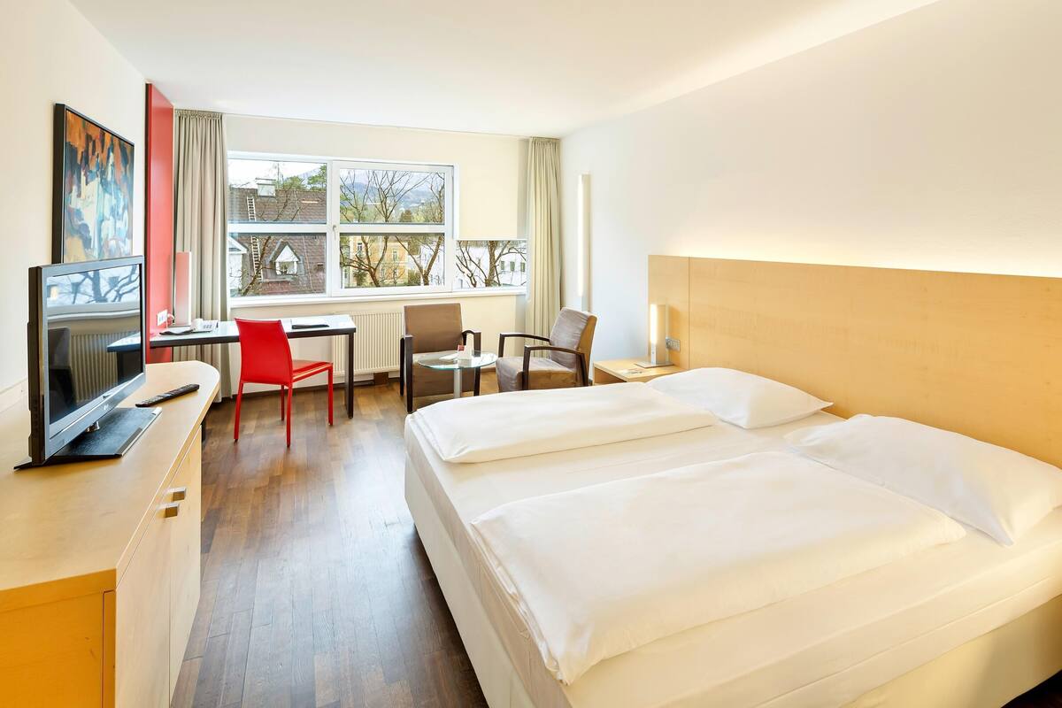 Una camera d'albergo con un letto, una scrivania con un computer, una sedia e una finestra vista edifici.