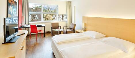 A hotel room with a bed, a desk with a computer, a chair, and a window with a view of buildings.