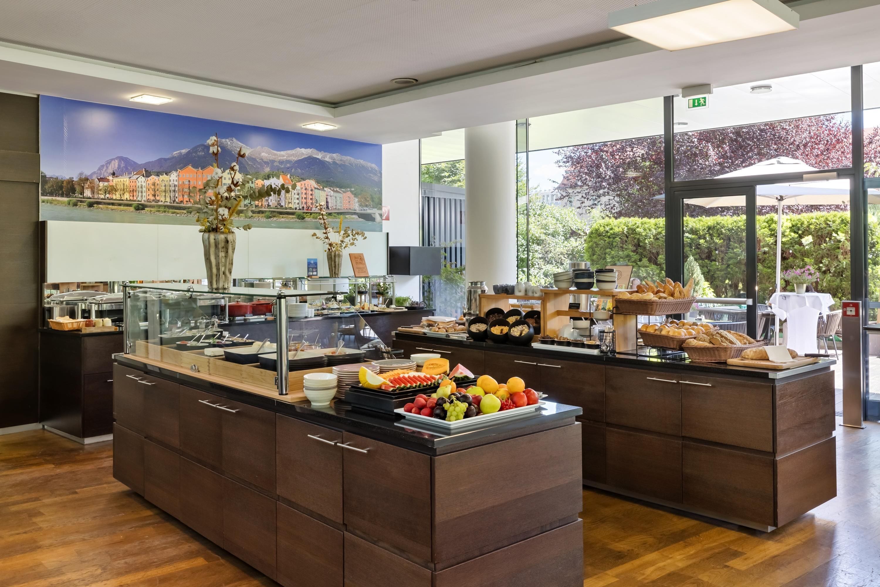 A buffet area with a variety of fruits and pastries, a large window with a view of buildings and mountains, and a modern interior design.