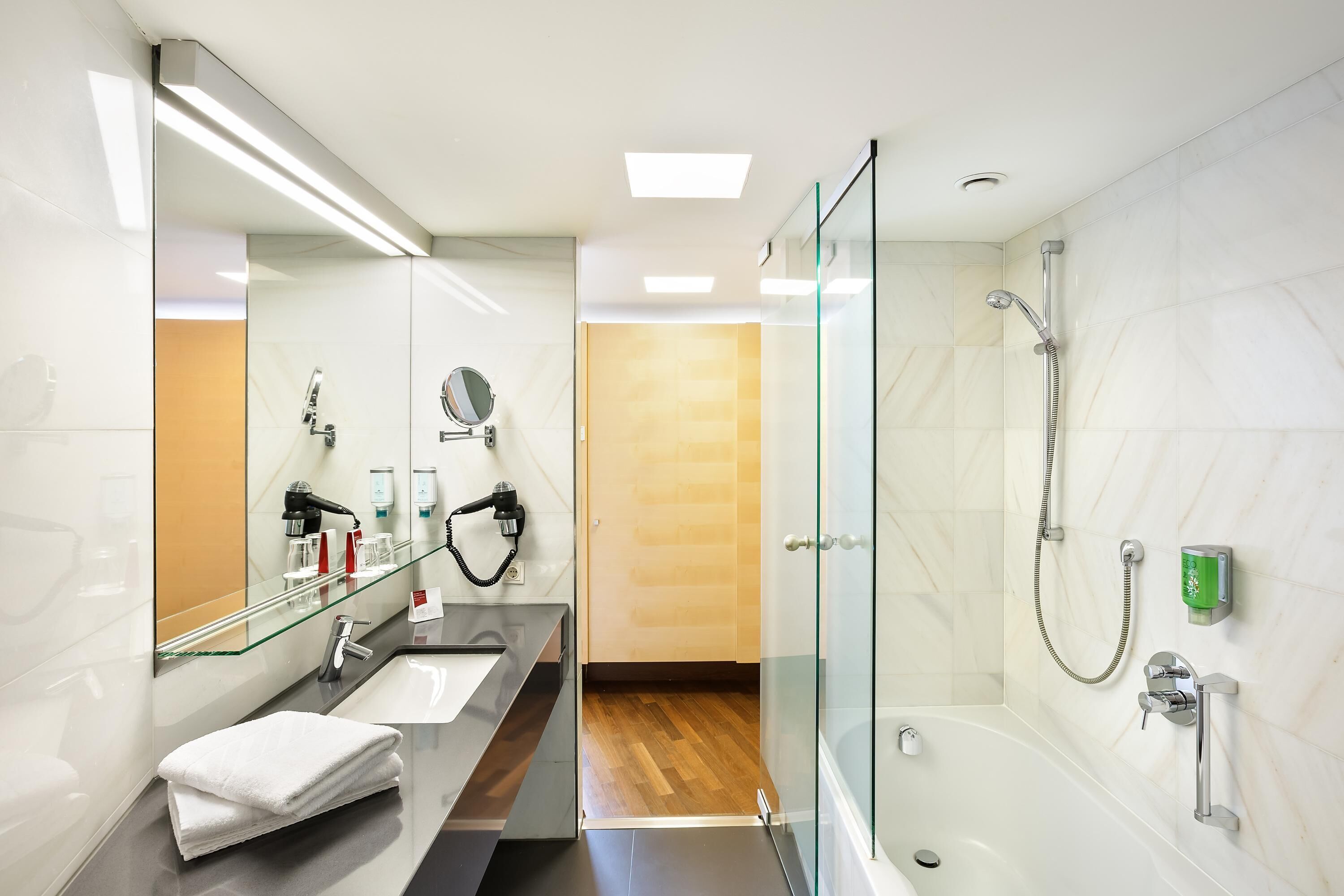 A modern bathroom with a glass shower enclosure, a large mirror, a vanity with a sink, and a towel stacked on the floor.