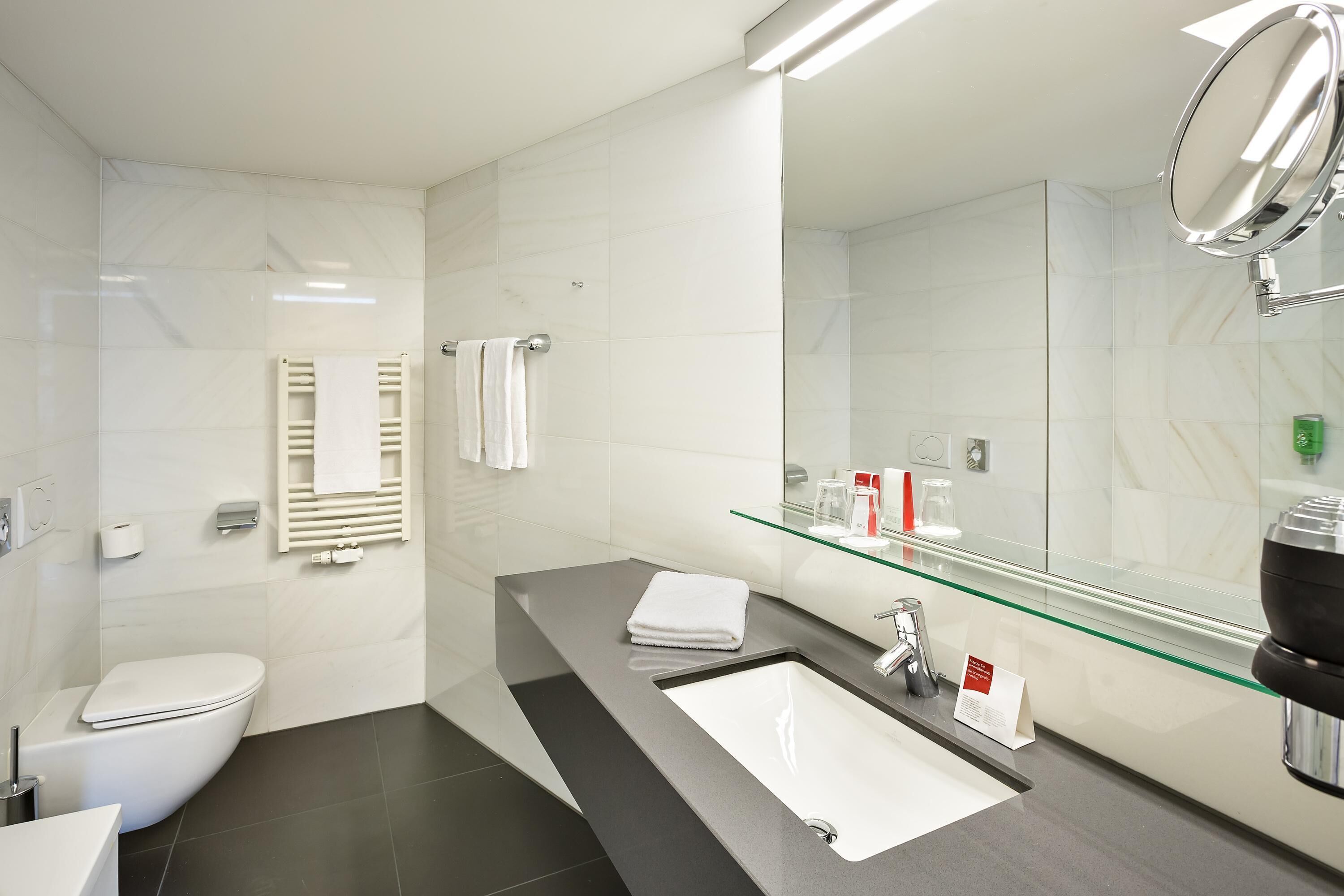 A modern bathroom with a white toilet, a glass shower enclosure, a black sink with a mirror, and a glass shelf with toiletries.