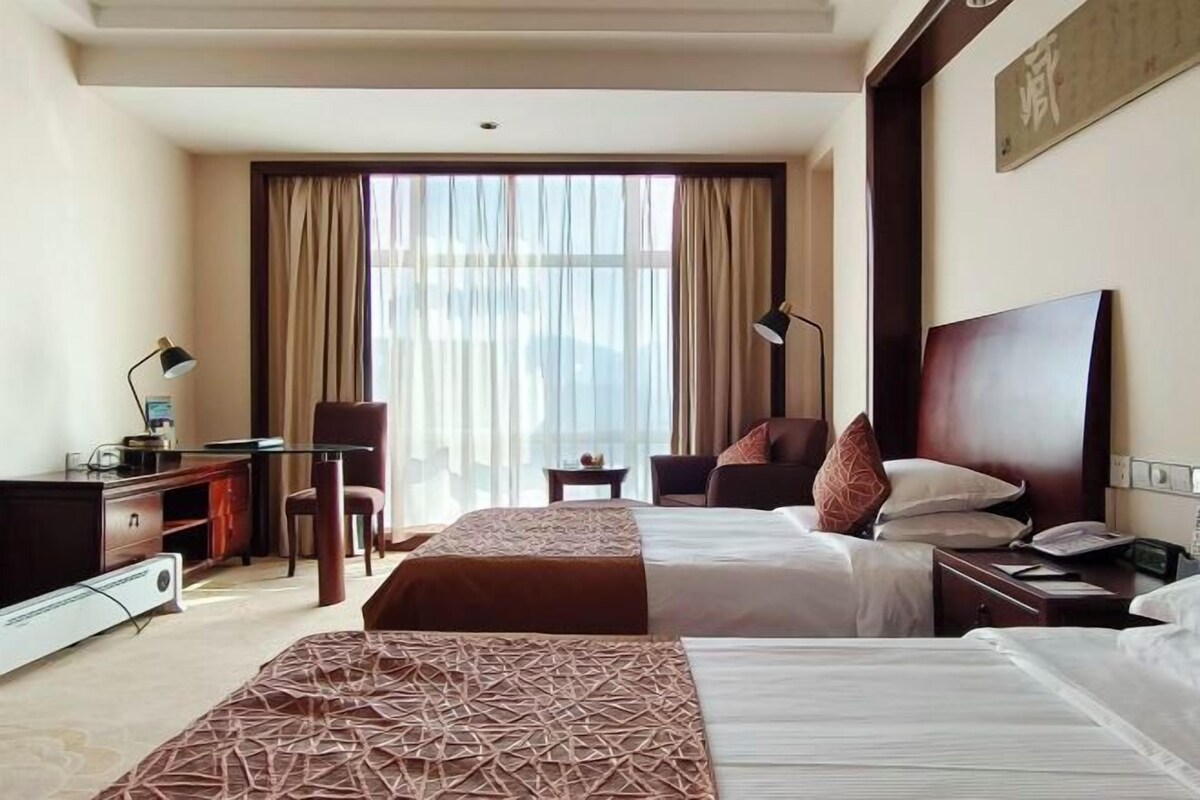 Ein Hotelzimmer mit zwei Betten, einem Schreibtisch, einem Stuhl, einer Lampe und einem großen Fenster mit Vorhängen.