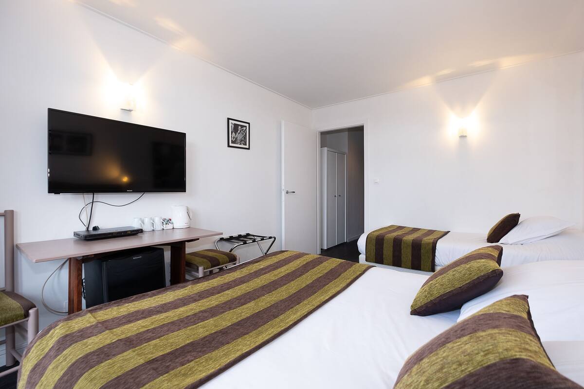 A hotel room with a flat-screen TV mounted on the wall, a desk with a computer and a coffee maker, a bed with striped bedding, and a door leading to another room.