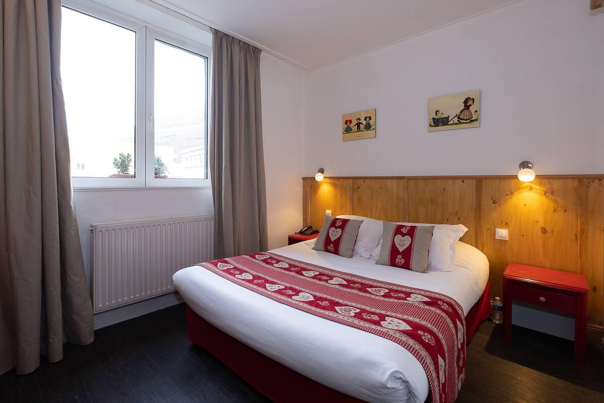 A bedroom with a large bed, a wooden headboard, two wall-mounted lights, a nightstand, a window with curtains, and two small wall art pieces.
