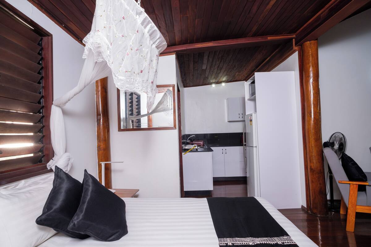 Une chambre à coucher avec un lit, des oreillers noirs, une couvre-lit blanche, un plafond en bois et une vue sur un coin cuisine.