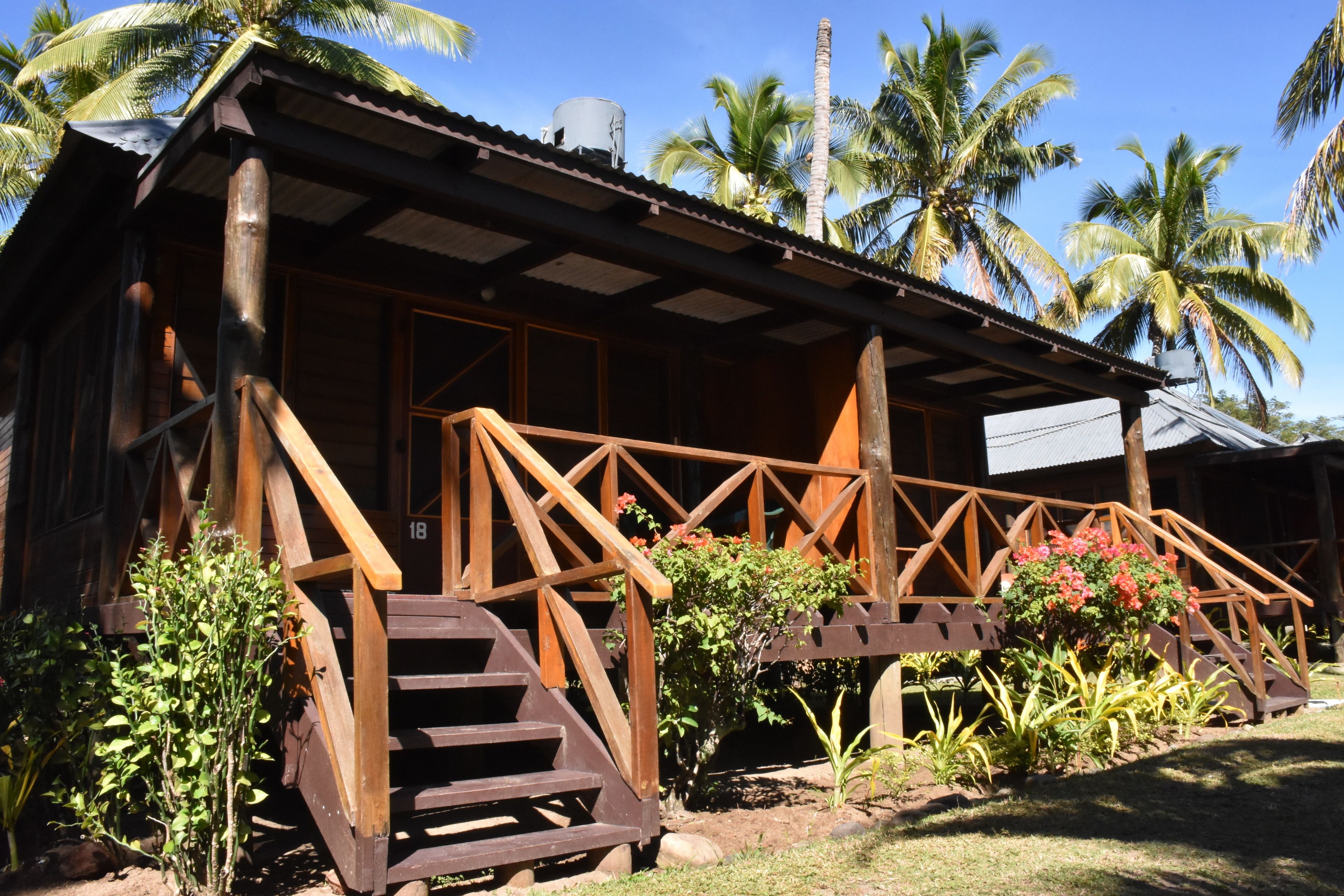 A wooden building with a porch and stairs, surrounded by tropical plants and palm trees.