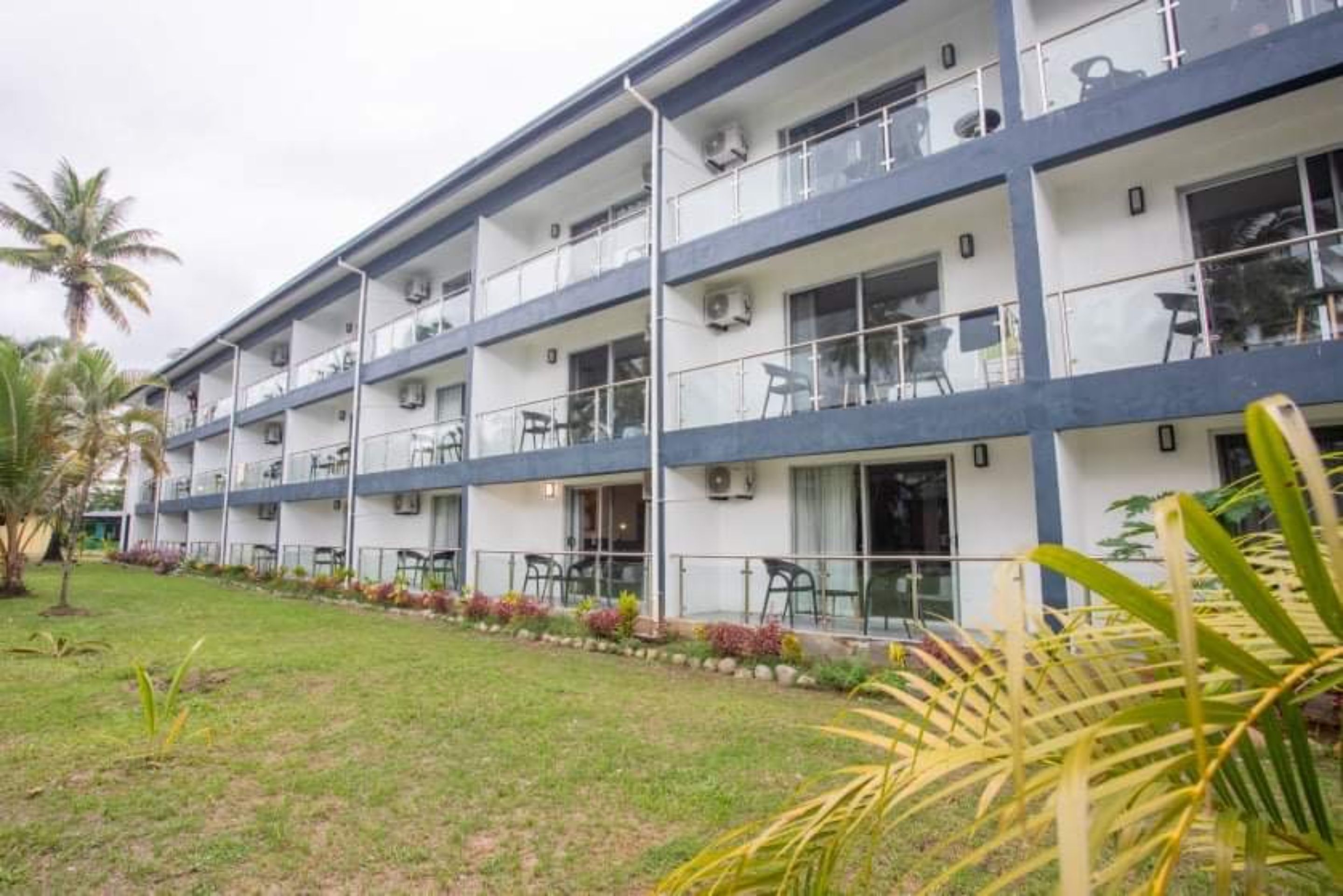 Un hôtel à plusieurs étages doté de balcons, de climatiseurs et d’une pelouse bien entretenue agrémentée de plantes tropicales.