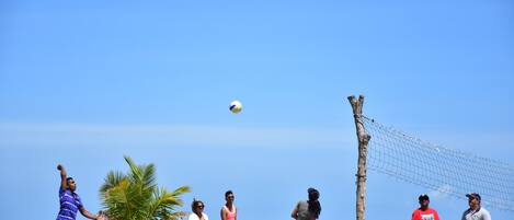Un grupo de personas jugando voleibol en la playa, con un bote y palmeras al fondo.