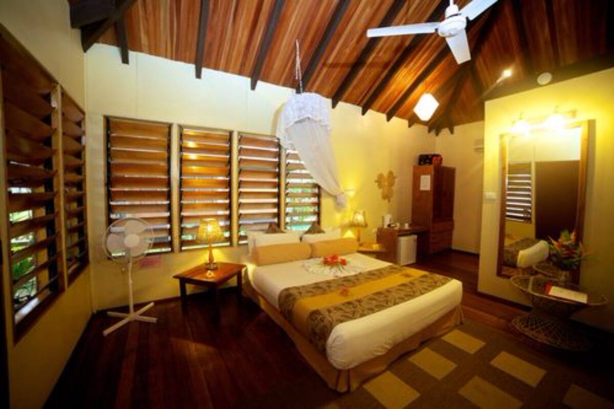 Une chambre d’hôtel avec un plafond en bois, un lit recouvert d’une courtepointe blanche et marron, une table de chevet avec une lampe, un ventilateur et un grand miroir agrémenté d’un arrangement floral.