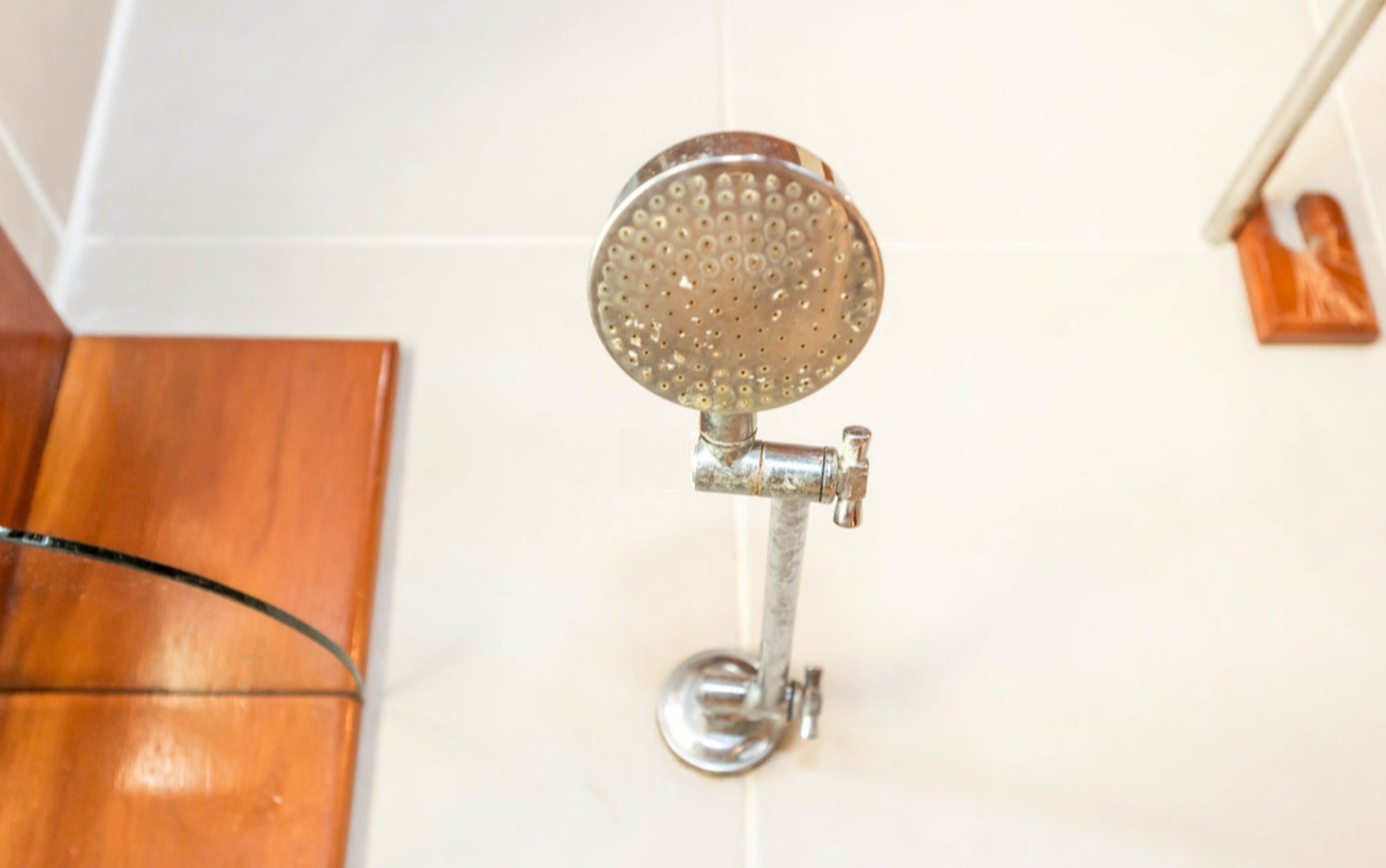 A showerhead mounted on a metal pipe in a bathroom with white tiled walls and a wooden floor.