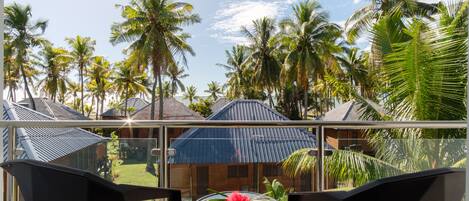 A balcony with a glass table and chairs, overlooking a tropical landscape with palm trees and a blue-roofed building.