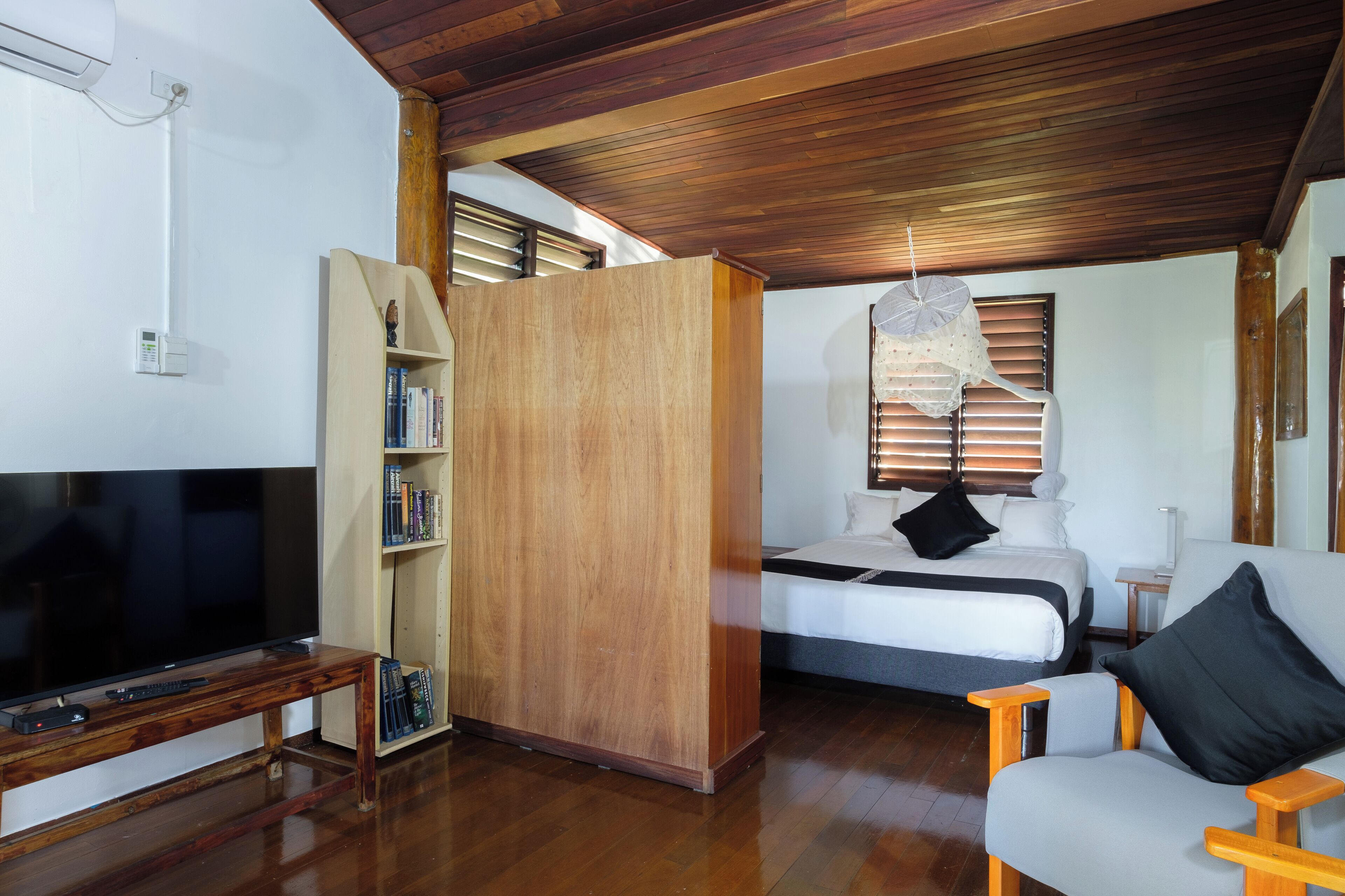 A room with a wooden ceiling, a bed with white bedding, a TV on a wooden stand, a bookshelf, and a window with blinds.