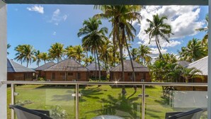 A balcony with a glass railing overlooking a lawn with palm trees and a building with a sloped roof.