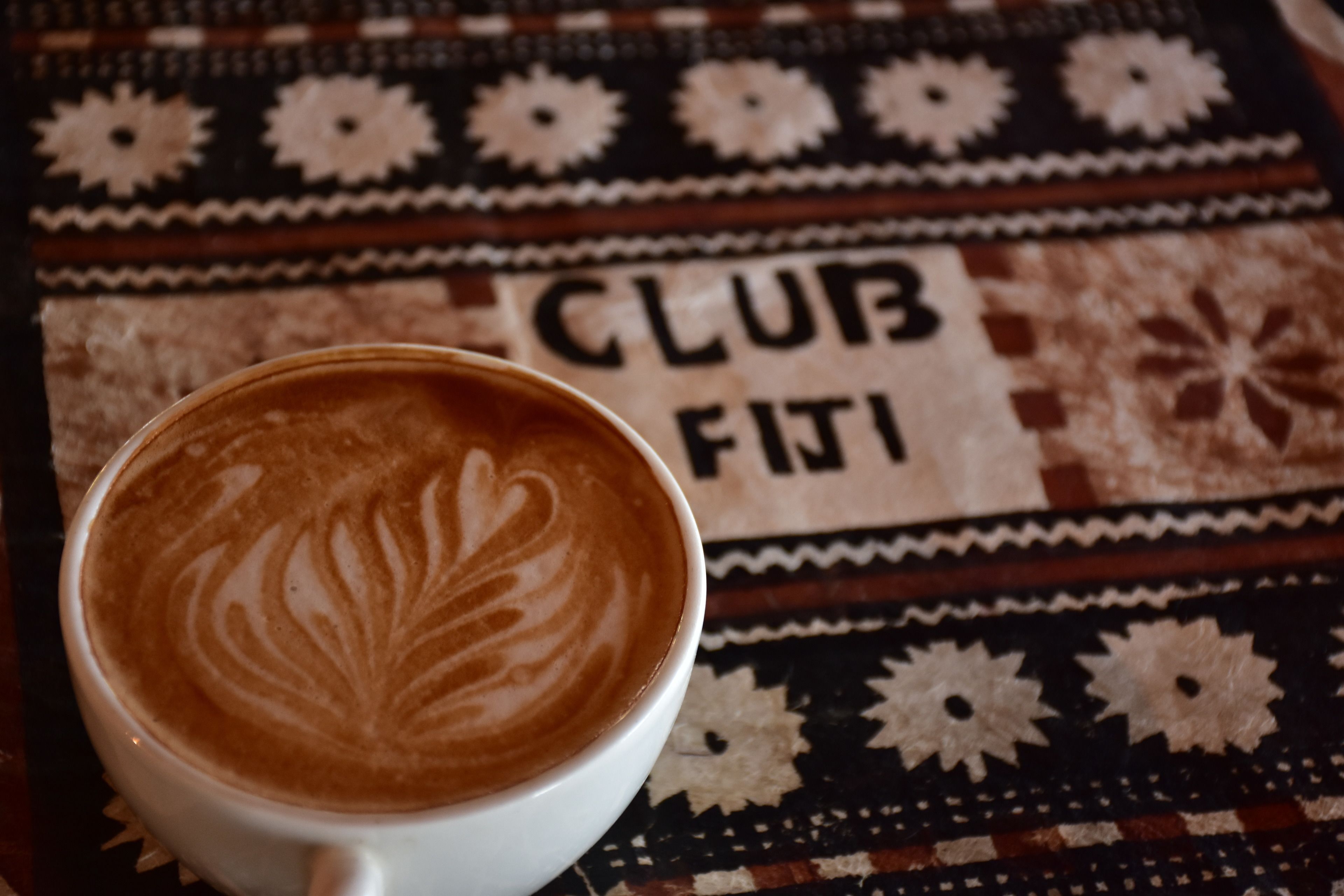 A cup of coffee with latte art on a patterned tablecloth with the text "CLUB FITI".
