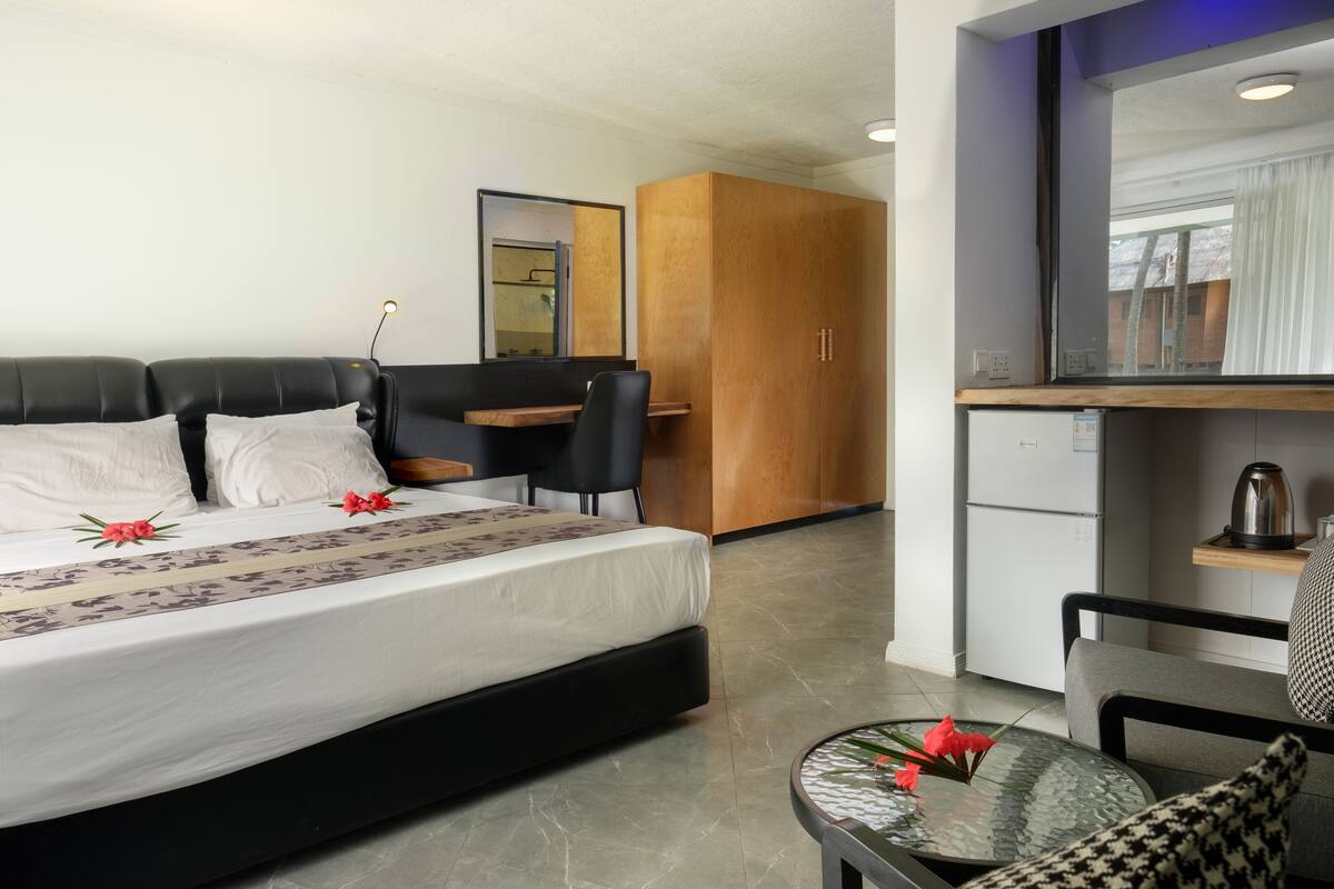 Une chambre d’hôtel moderne comprenant un lit, une petite table avec un vase de fleurs, un coin salon avec un fauteuil et une kitchenette.
