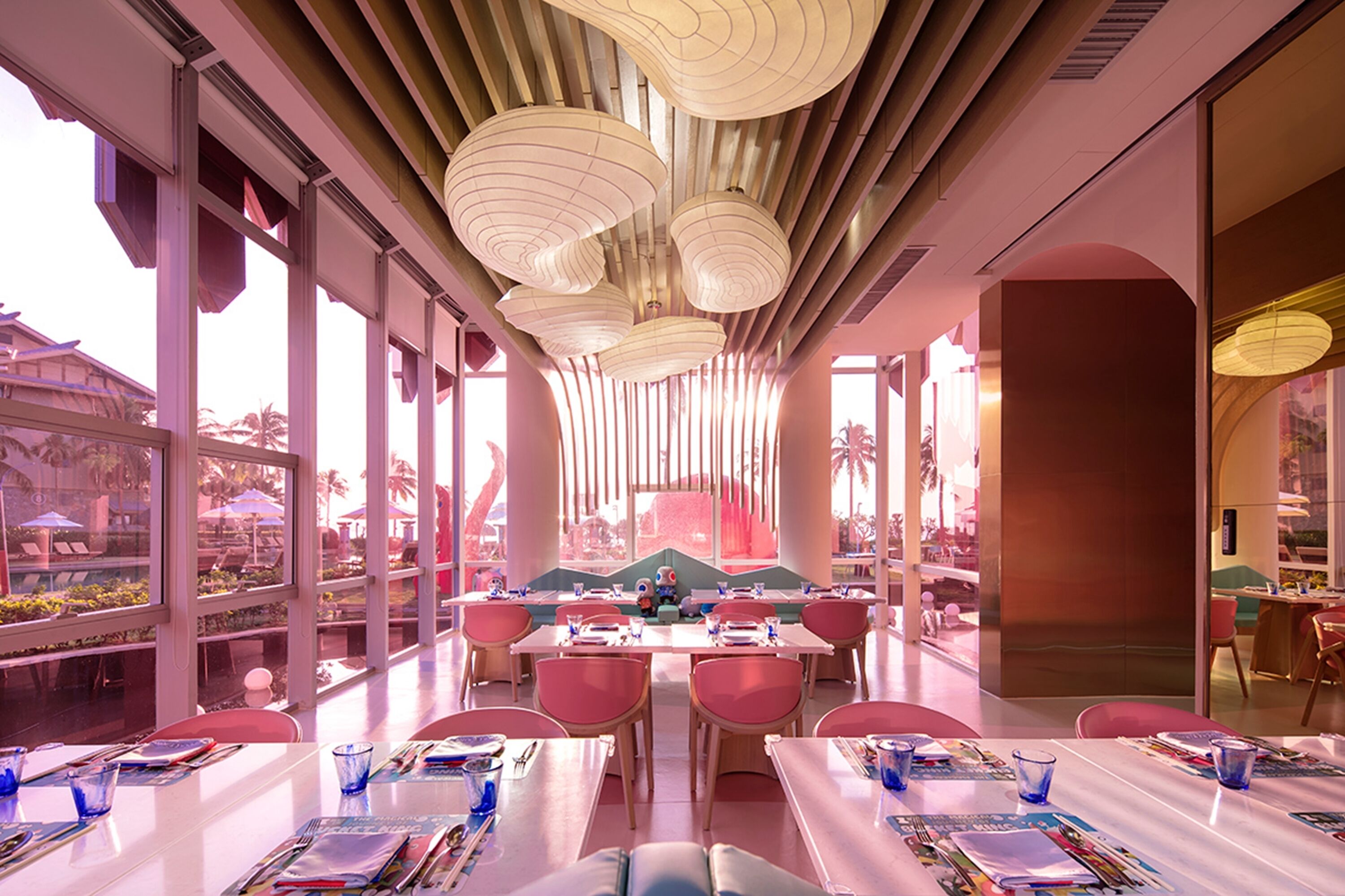 Ein Restaurant mit hohen Decken, moderner Pendelleuchten und großen Fenstern, die Ausblicke ins Freie bieten. Die Tische sind mit blauem Glas und Speisekarten gedeckt.