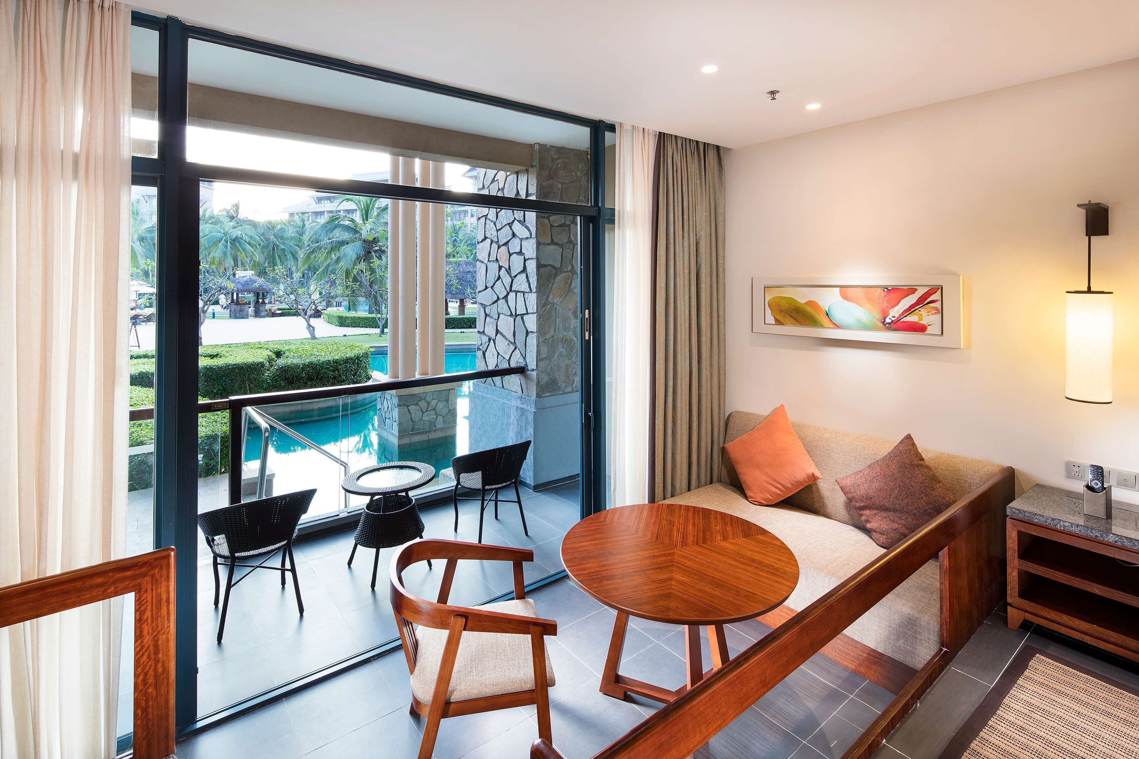 A modern hotel room with a balcony, a sofa, a round wooden table, and a painting on the wall.
