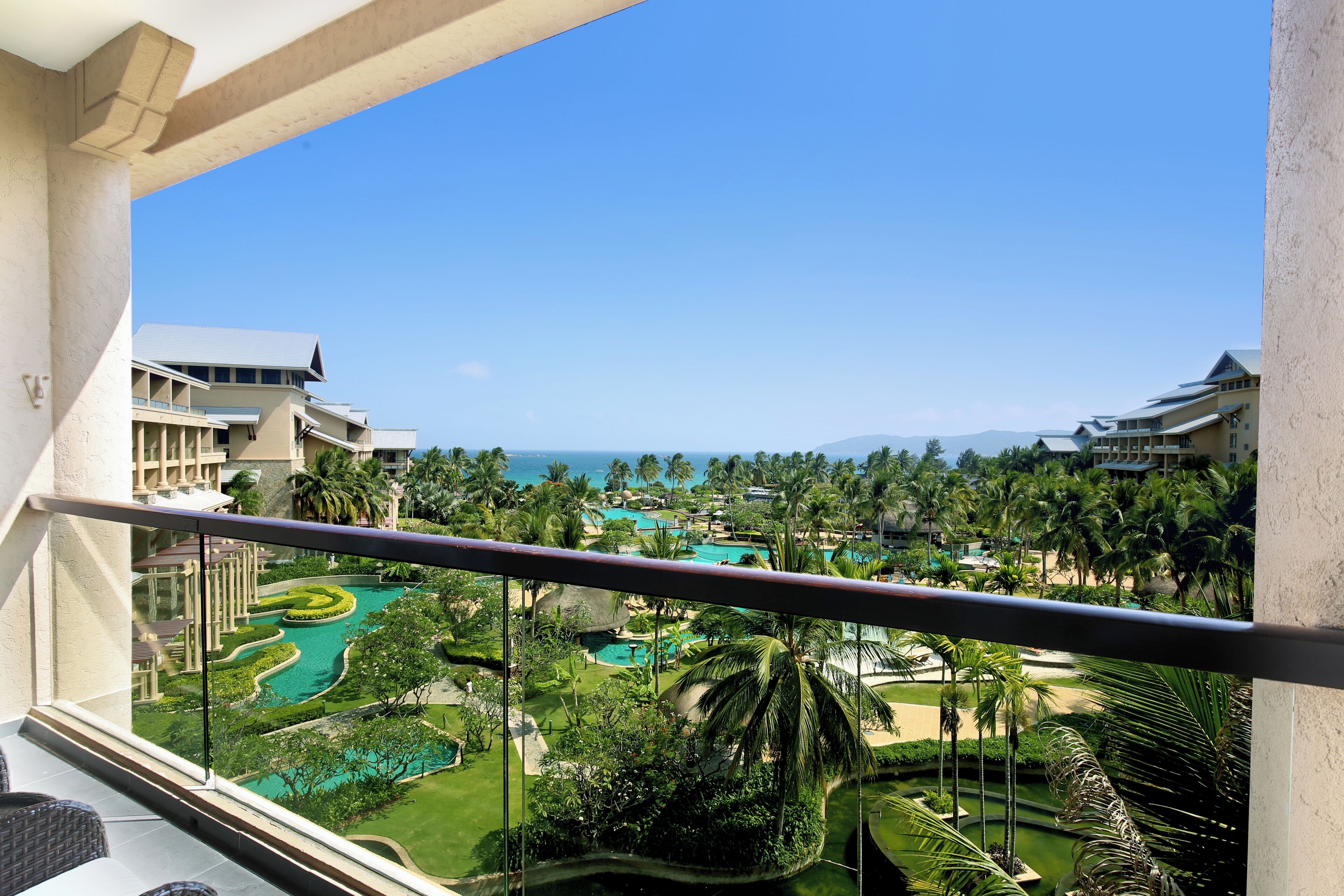 A resort balcony with a view of a landscaped pool area, palm trees, and a distant ocean.